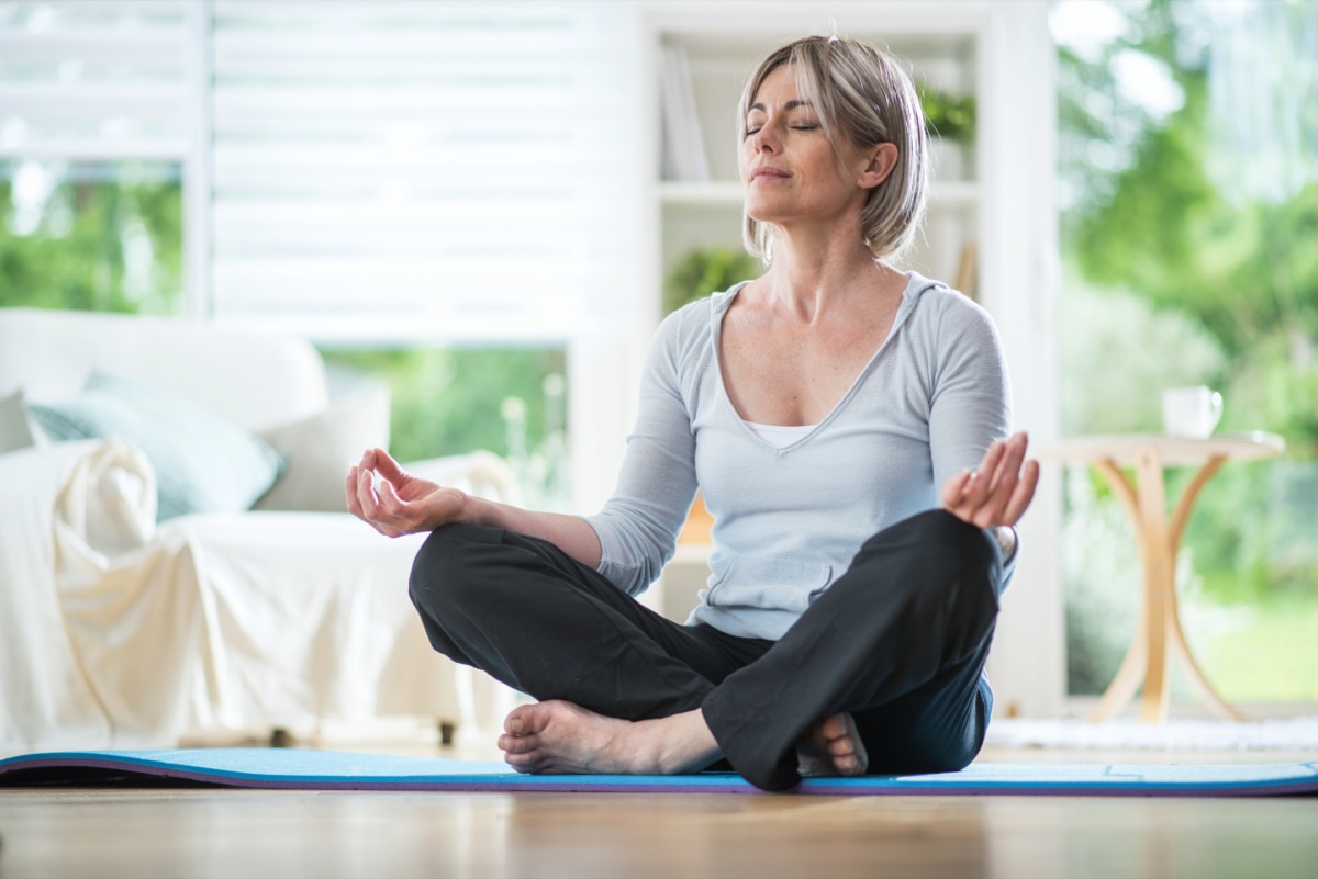 Middle aged woman sitting in lotus position on a carpet in his living room. her eyes are closed. she is in the foreground