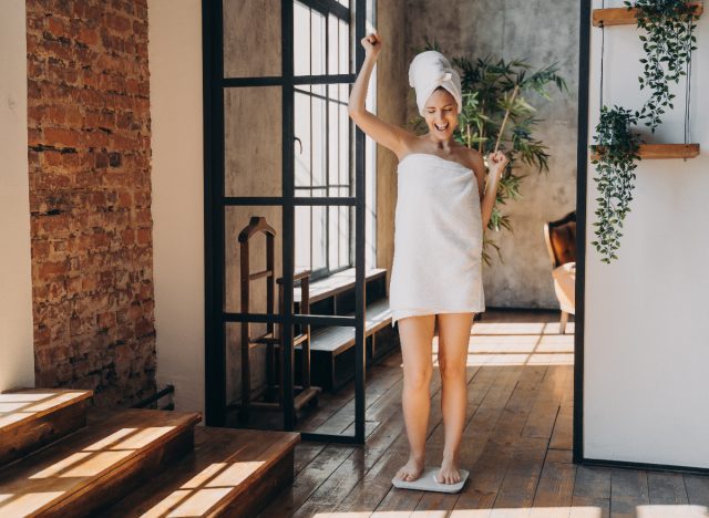 happy woman in towel weighing herself after shower, concept of how to get the scale moving after a weight loss plateau