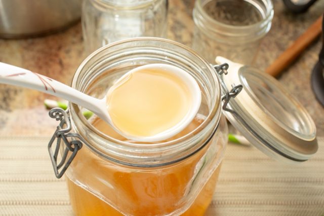 A view of a ladle scooping some bone broth from a large glass jar.